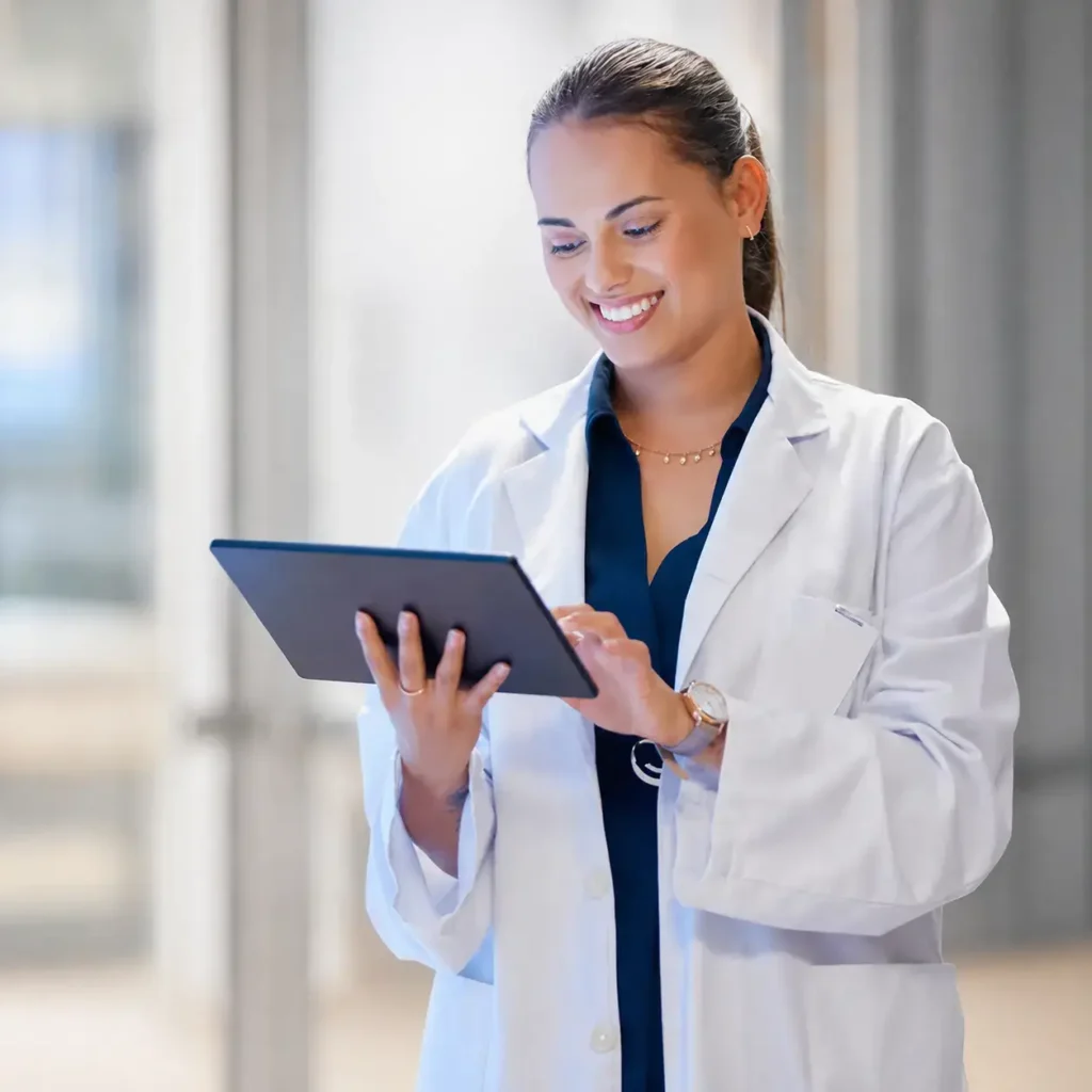 Healthcare professional in a white lab coat over navy medical scrubs using a tablet in a modern private practice clinic.